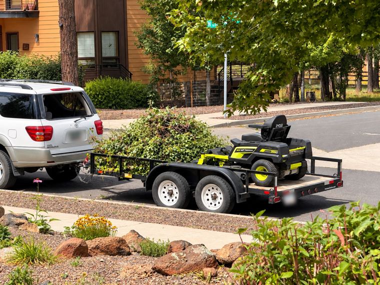 Tandem trailer loaded with a mound of cleared brush and a zero turn mower